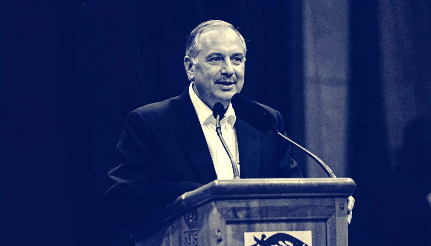 Duotone photograph of a speaker at a podium, deep navy and warm ivory tones