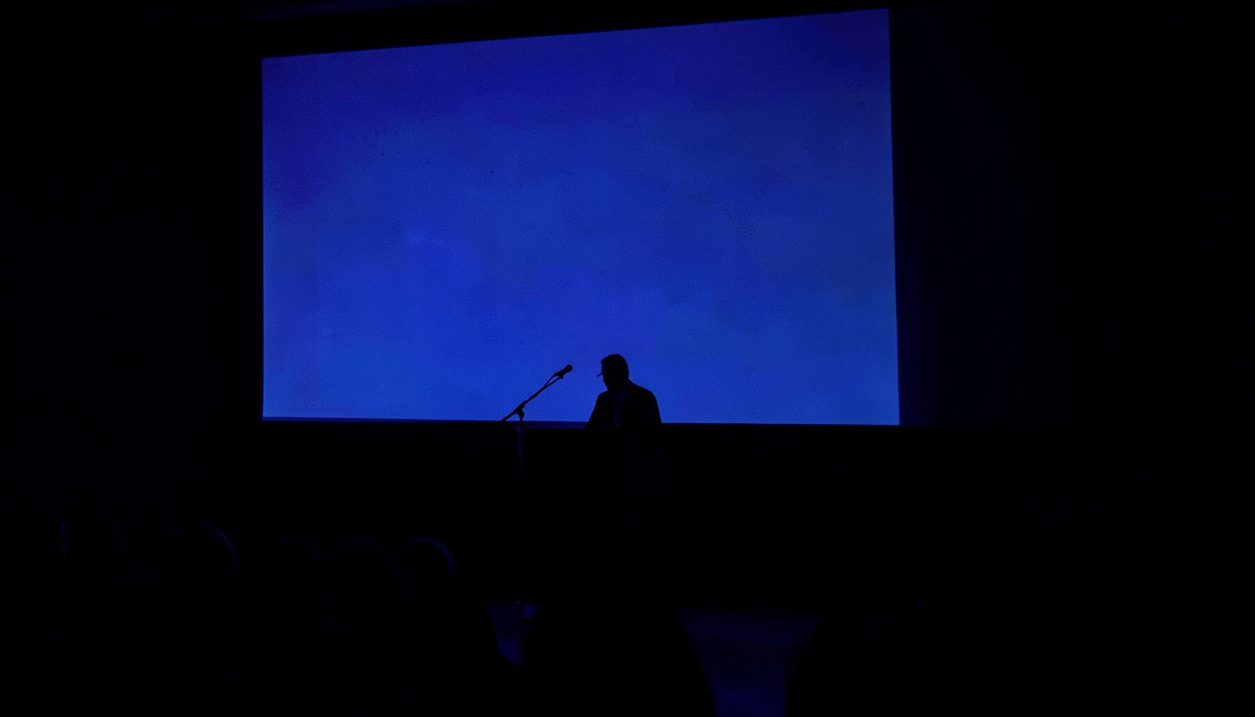 A speaker at a podium silhouetted against a large projection screen in a darkened auditorium, deep indigo duotone