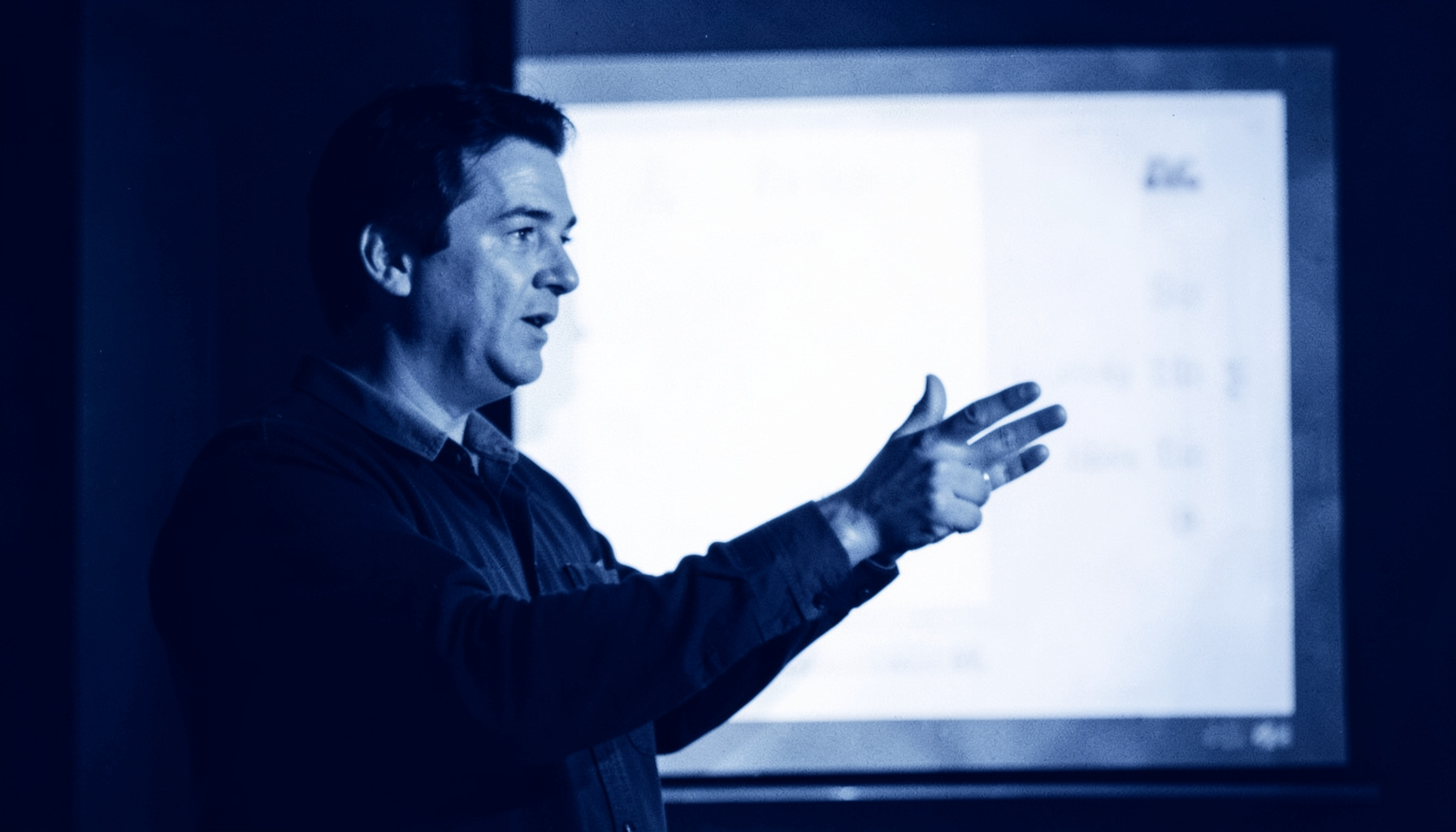 A person gesturing at a presentation screen in a conference setting, deep navy and soft white duotone